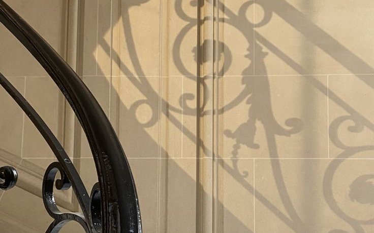 A wrought iron banister casting its shadow on a stone stairwell wall